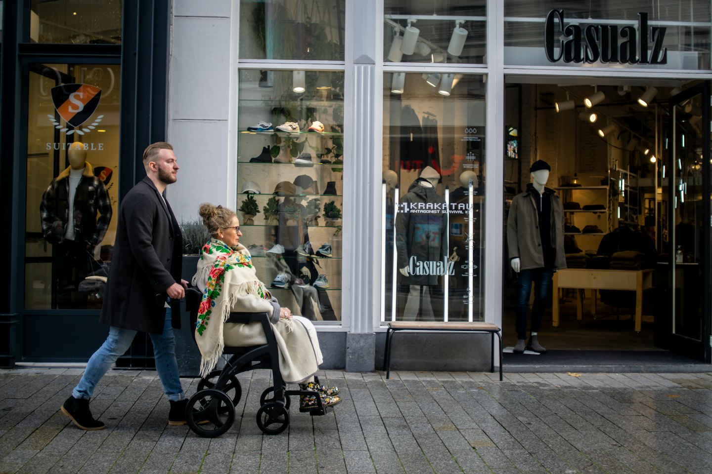 Man pushes woman in wheelchair past city storefronts.